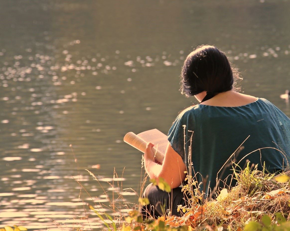 Looking at a dark haired girl in a blue shirt reading a book by a lake. Her back is to us.