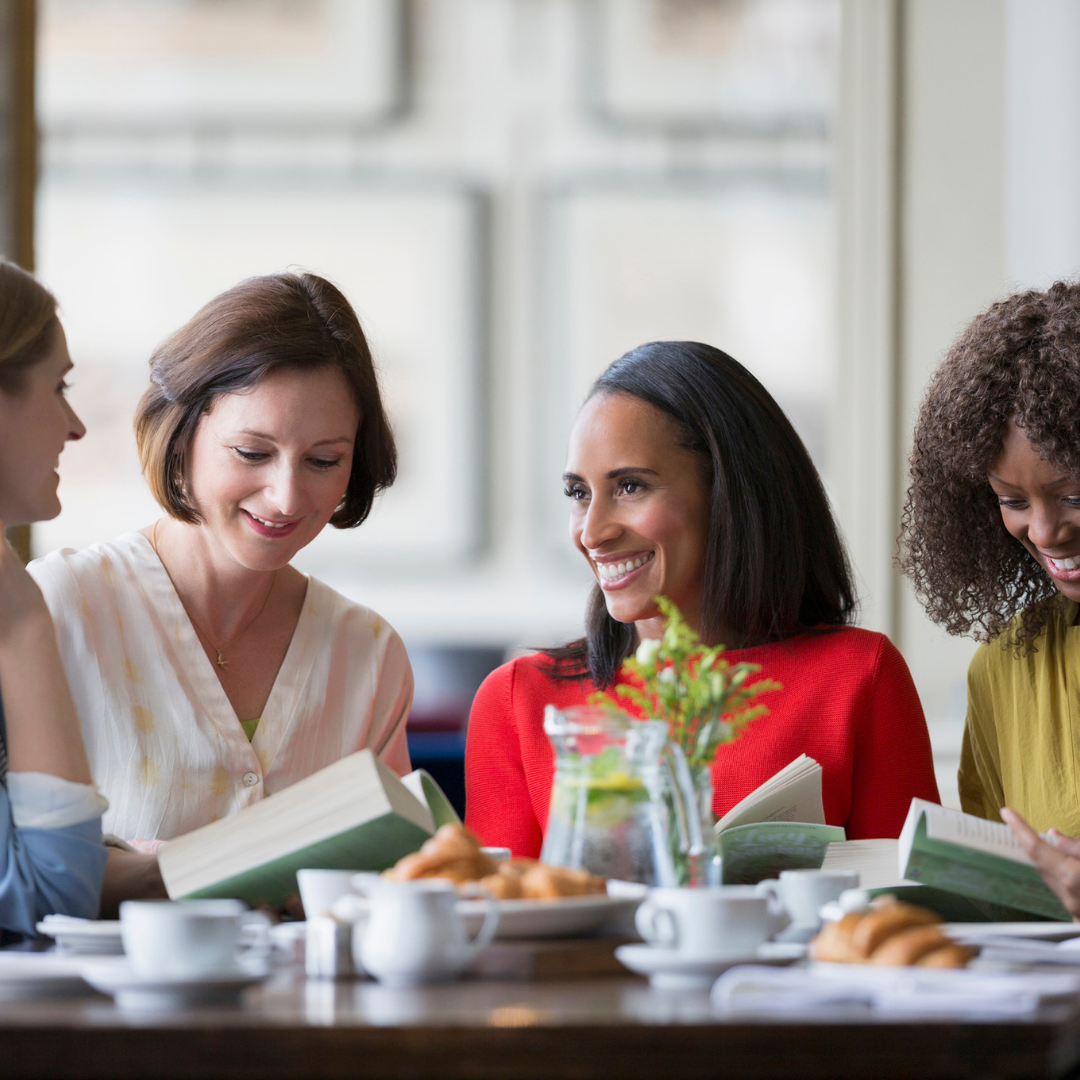 women at a table with dishes and open books, talking. 