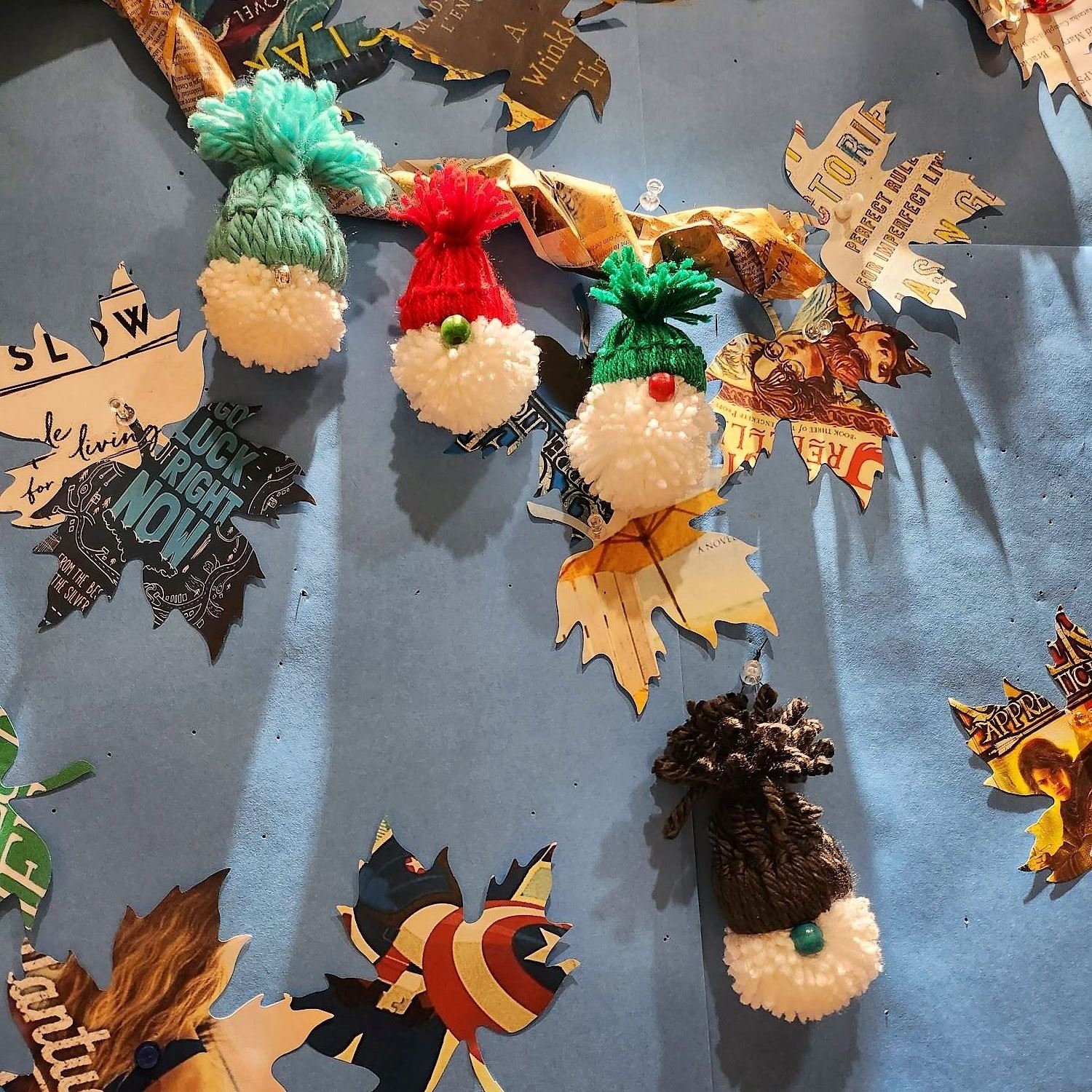 4 white pom-pom yarn gnomes hanging on a brown newspaper tree, all on a blue background.