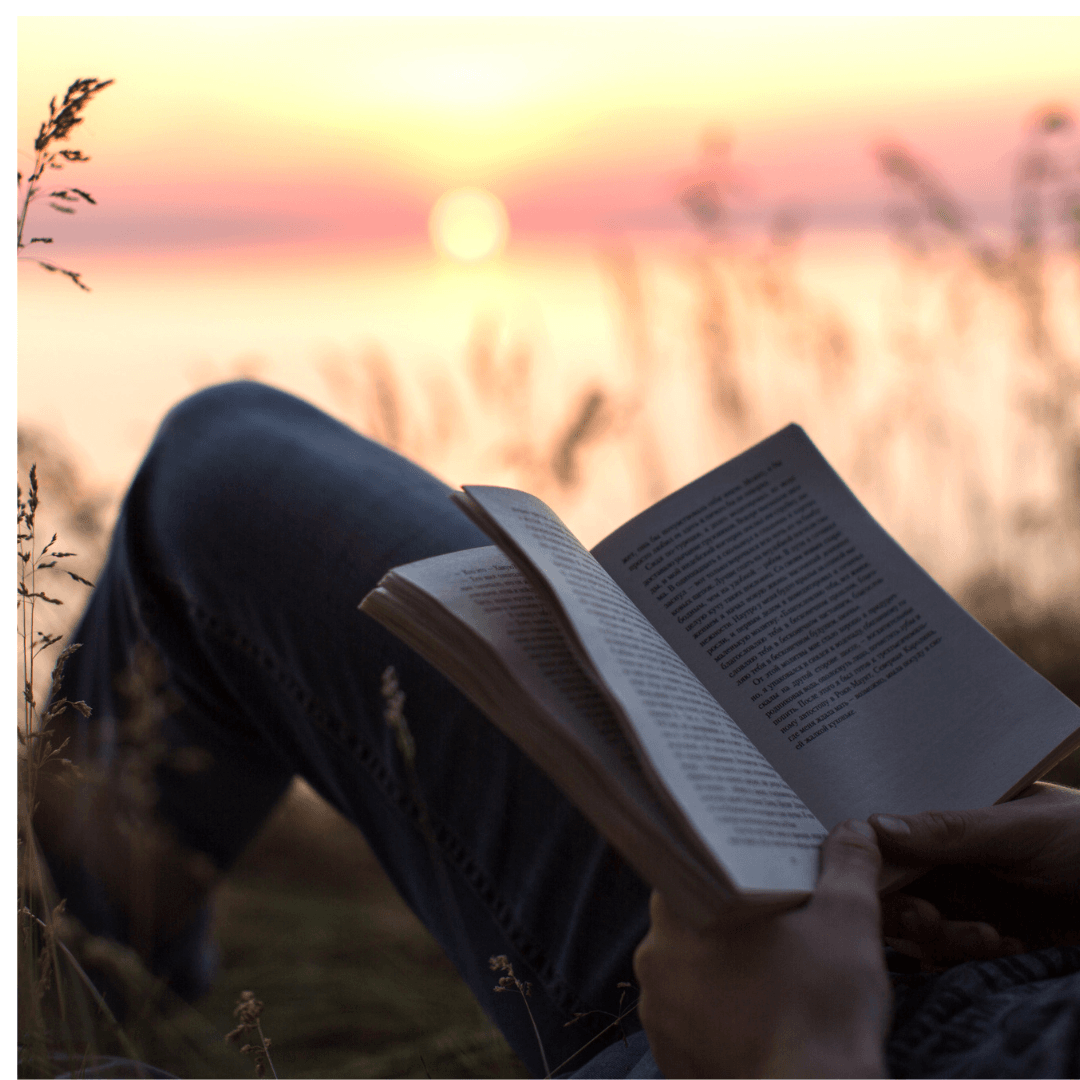 Reader laying down with a book on their knees in tall grass with sunset over water