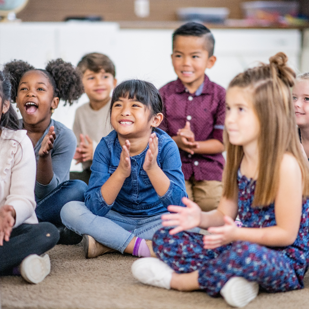 Photo of kids clapping while sitting on the floor, looking at someone